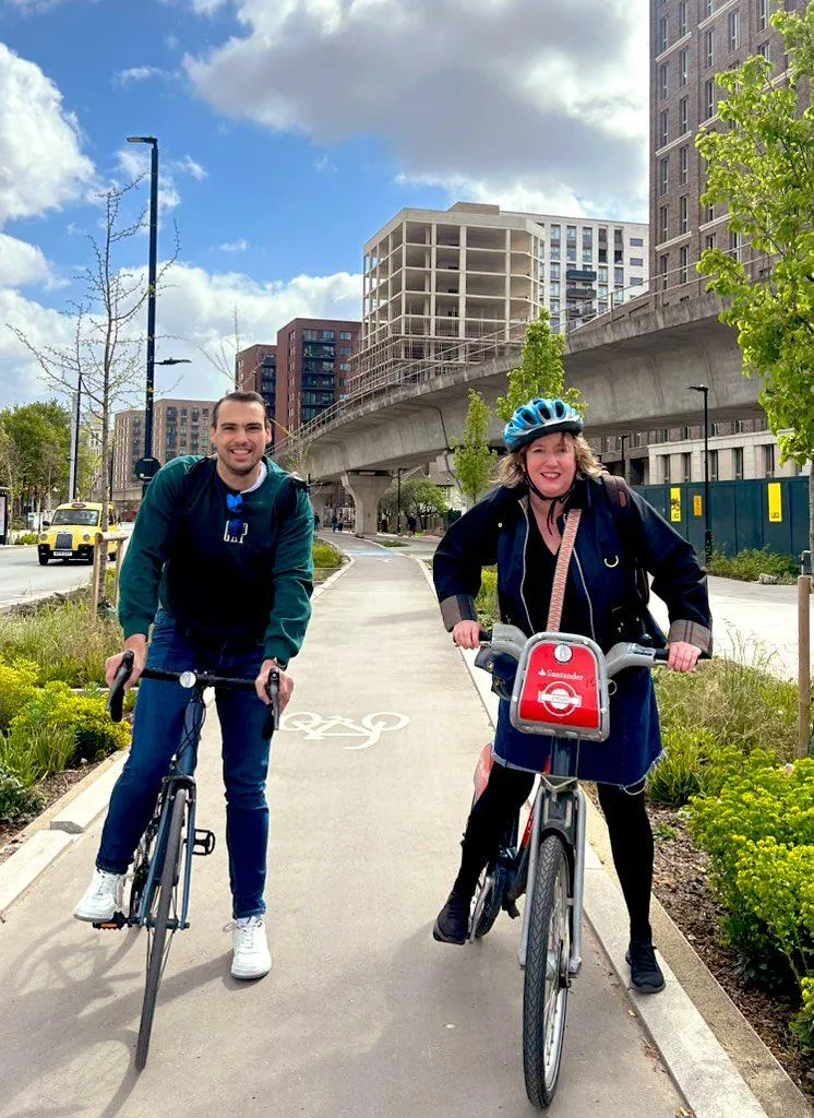 John Morris and Carla cycling together along the Royal Docks Corridor, with the cable car and docklands towers behind them